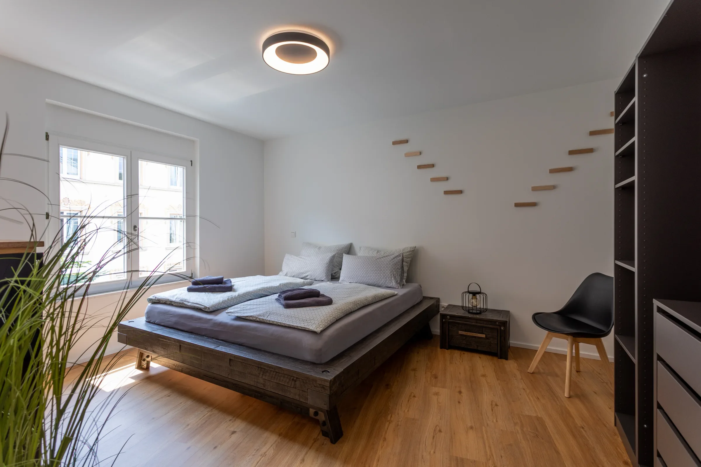 Hell eingerichtetes Schlafzimmer mit niedrigem Holzbett, Fenster, runder Deckenleuchte und offenem Regal