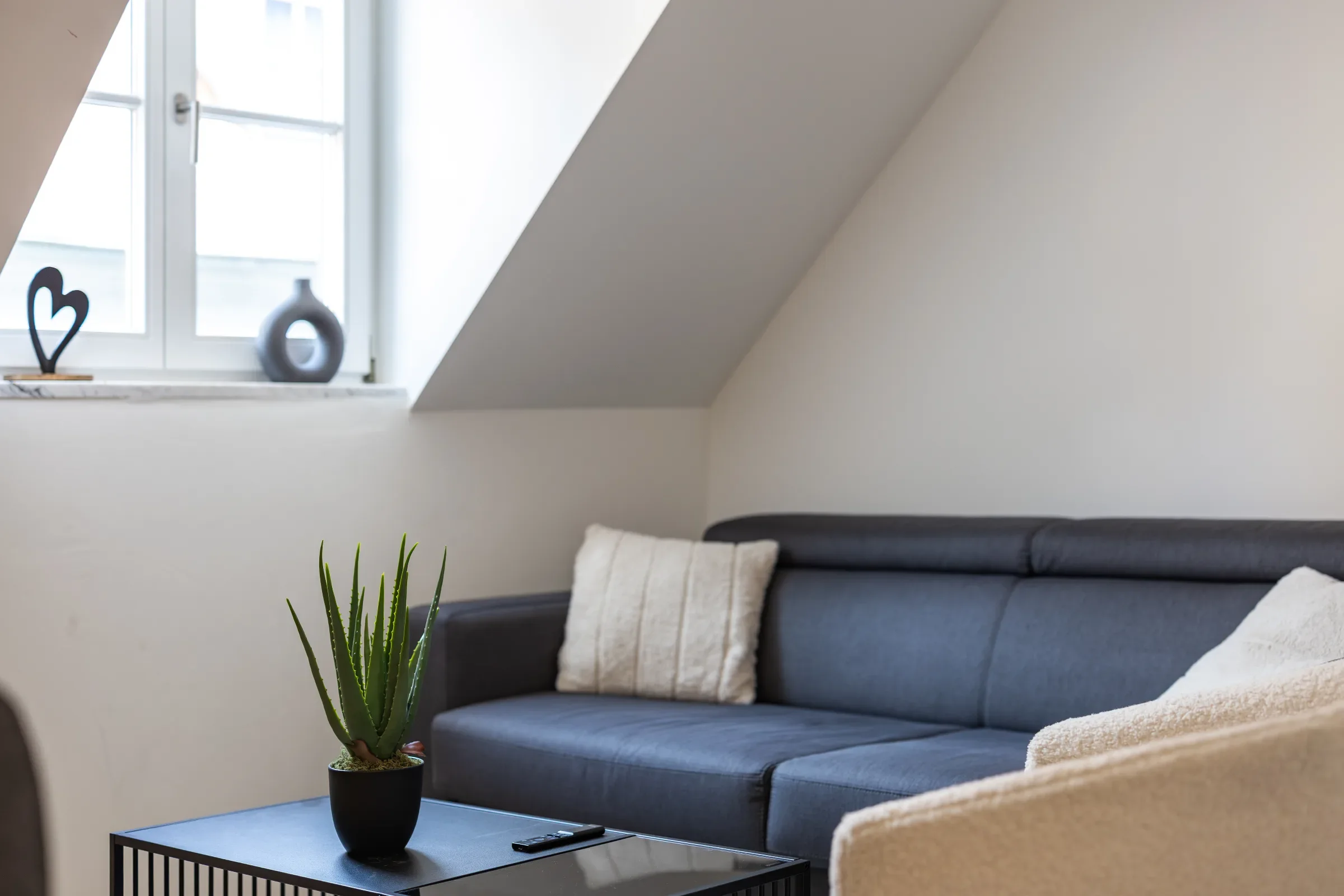 Dunkelgraues Sofa unter Dachschräge, Fensterbank mit Deko, kleiner Couchtisch mit Topfpflanze im Vordergrund.