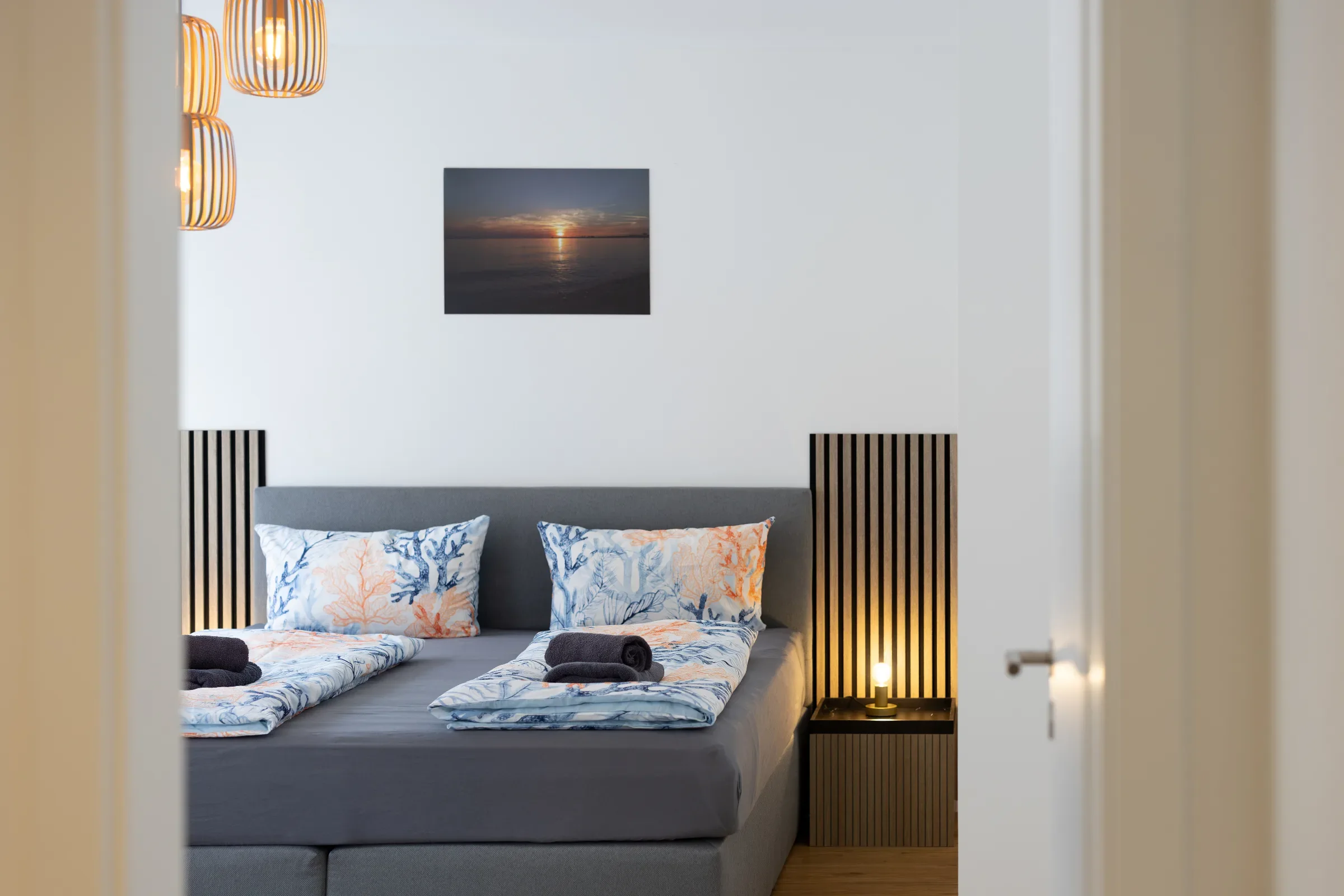 Blick durch offene Tür auf Doppelbett mit grauem Kopfteil, gemusterten Kissen und kleinem Wandbild
